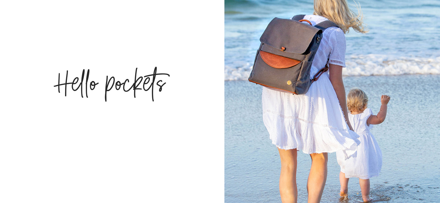 Image of a mom wearing a grey canvas DUO Backpack, walking her toddler at the beach shoreline, with text overlay "Hello Pockets"