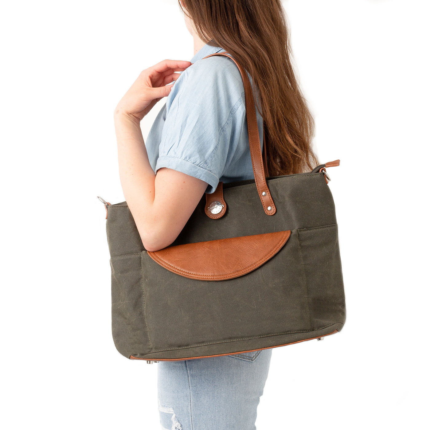 Woman in a blue blouse and jeans standing sideways and carrying a forest green waxed canvas carryall tote with caramel brown vegan leather accents, positioned against a white background.