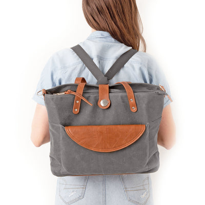 Woman in blue blouse and jeans standing backwards against a white wall and wearing a grey tote bag as a backpack with caramel brown vegan leather accents.