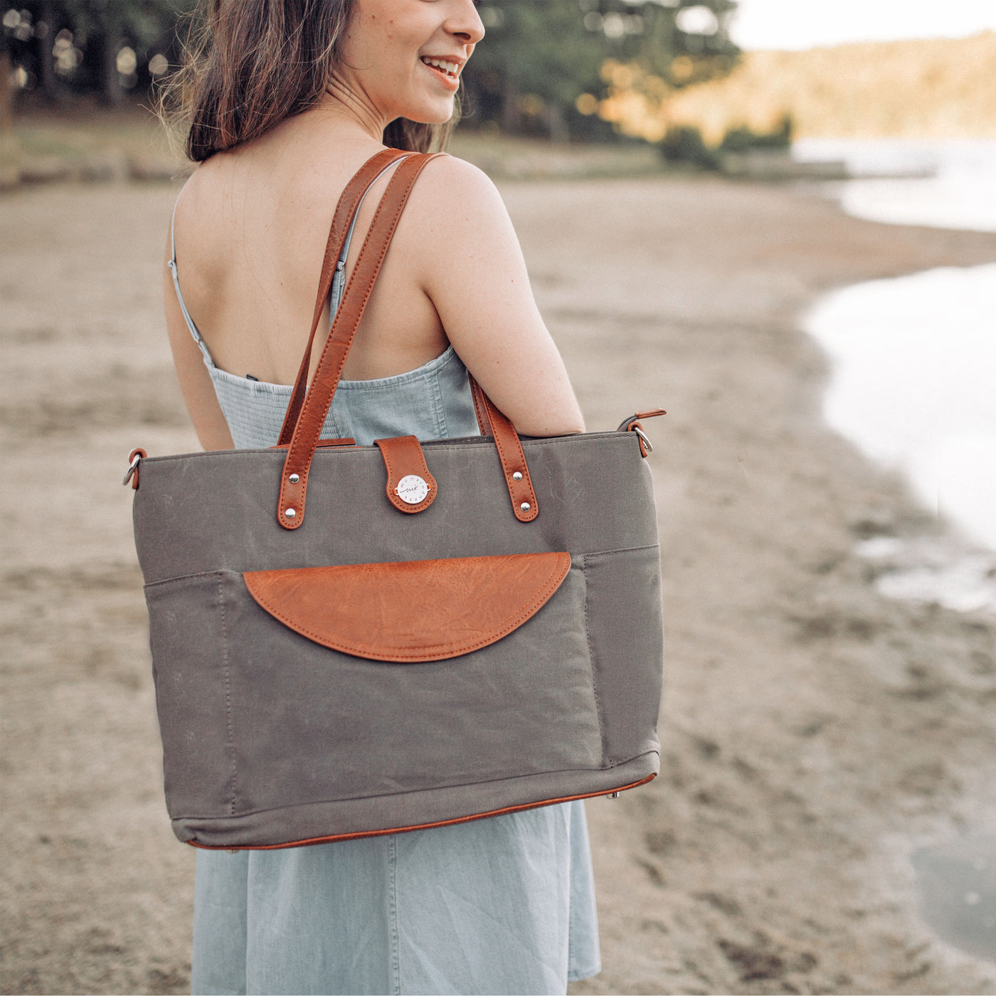 A woman in a denim dress standing outside by a lakeshore carrying a grey canvas carryall tote bag with caramel brown vegan leather accents. 