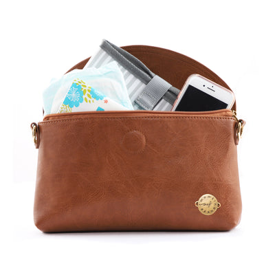 Vegan leather clutch shown with included folded changing pad and additional mom & baby items including a cell phone, baby diapers and wipes.  Brown vegan leather clutch has antique bronze metal Momkindness logo and D-rings on side to attach included adjustable vegan leather crossbody strap.