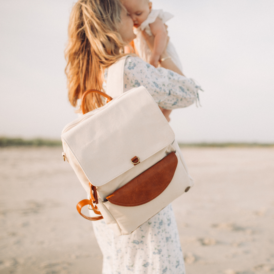 Women standing at water view wearing white colored backpack with brown vegan leather accents on her back. Beach surroundings with her baby held close.  