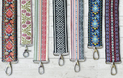A group of 7 brightly colored, patterned messenger-style bag straps with silver clasps, on a white wood background.
