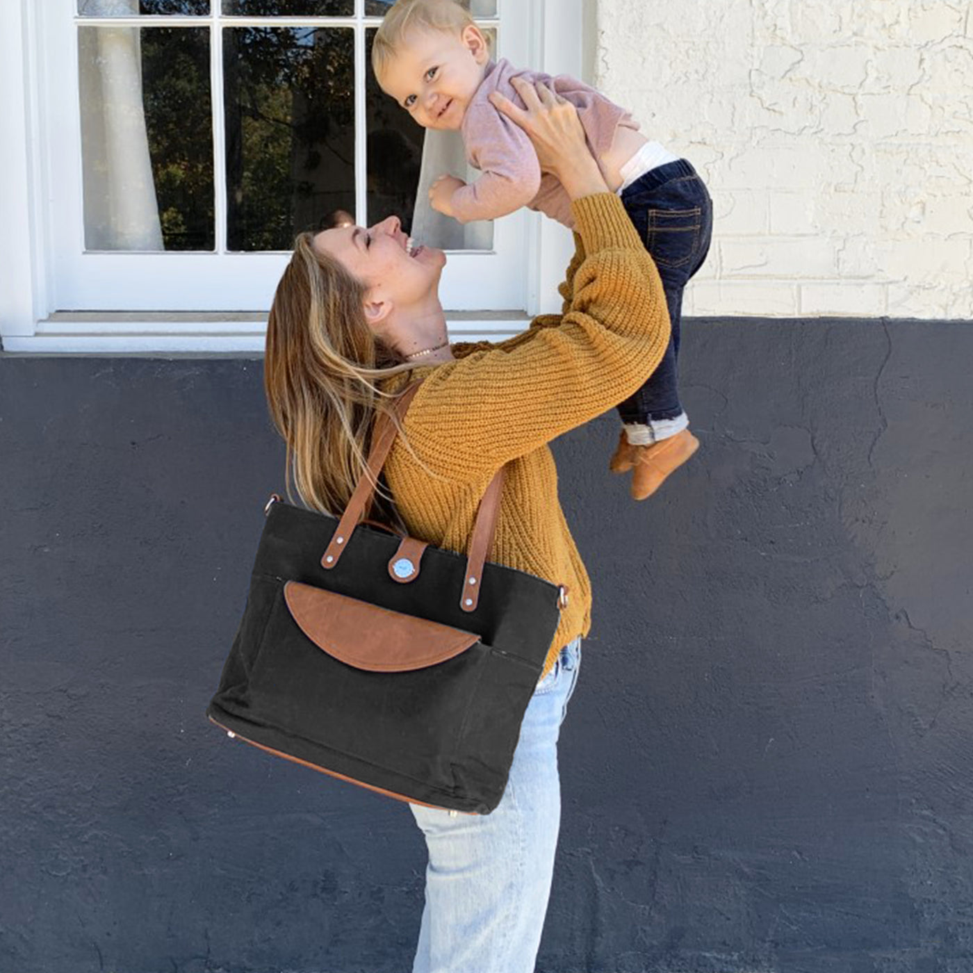Mom standing sideways holding baby in the air, wearing a black waxed canvas tote with brown vegan leather accents on her shoulder. 