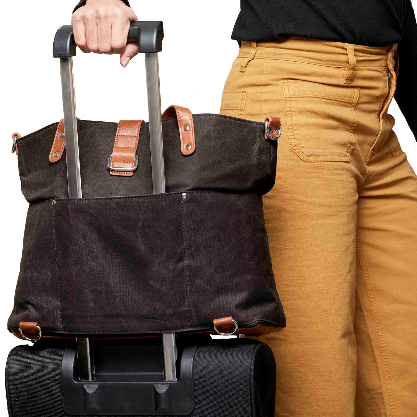 Woman walking with black waxed canvas tote attached over handle of rolling suitcase via trolley sleeve on backside of tote.