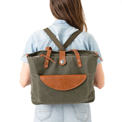A mom in a blue blouse and jeans standing backwards wearing a forest green waxed canvas backpack with brown vegan leather accents, all on a white background.