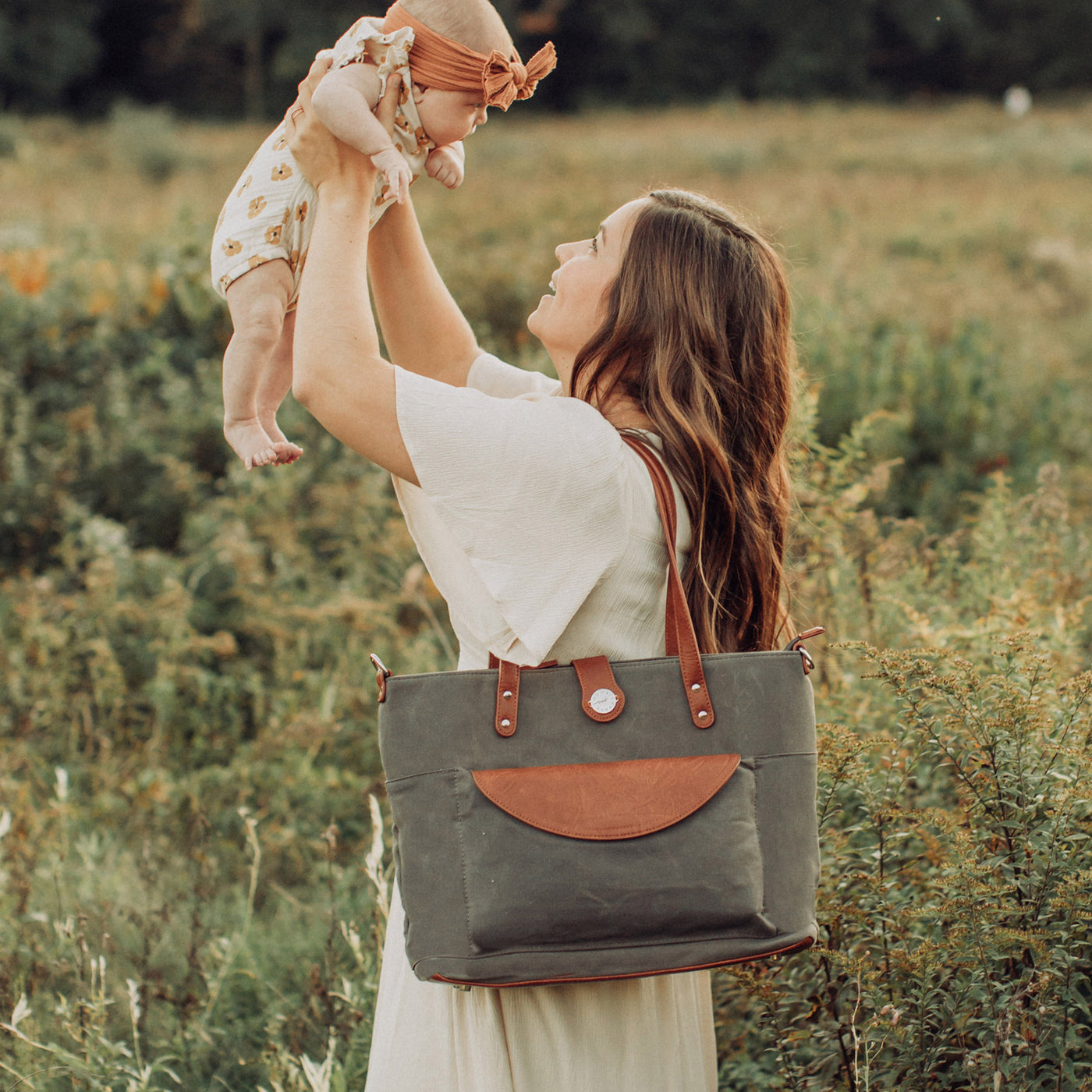 A mom holding her baby girl in the air, standing in a grass field, wearing a forest green waxed canvas CarryAll Tote with caramel brown vegan leather accents on her shoulder.