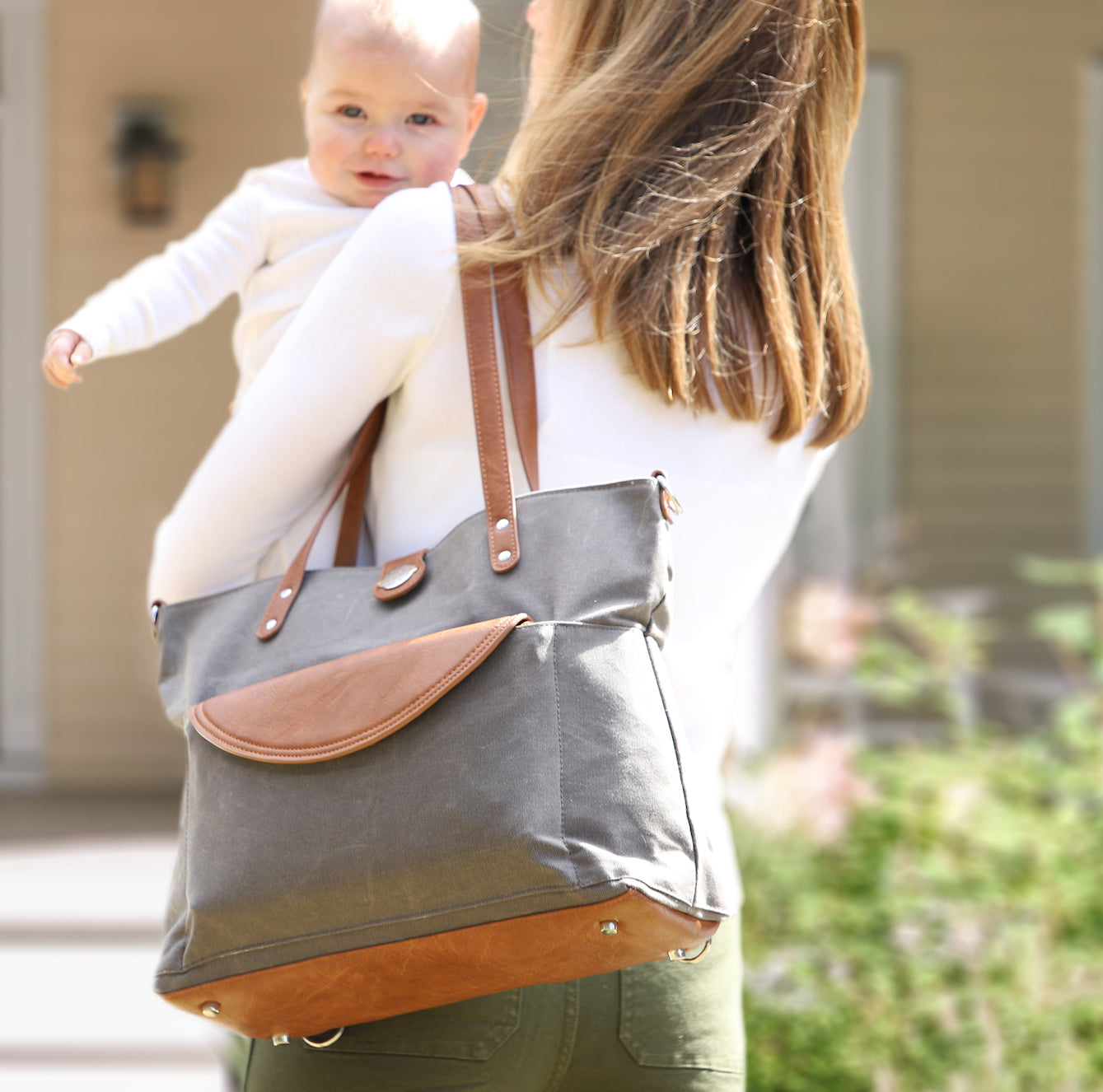 Mom outside front door carrying baby and wearing a grey waxed canvas CarryAll Tote with brown vegan leather accents on her shoulder. 