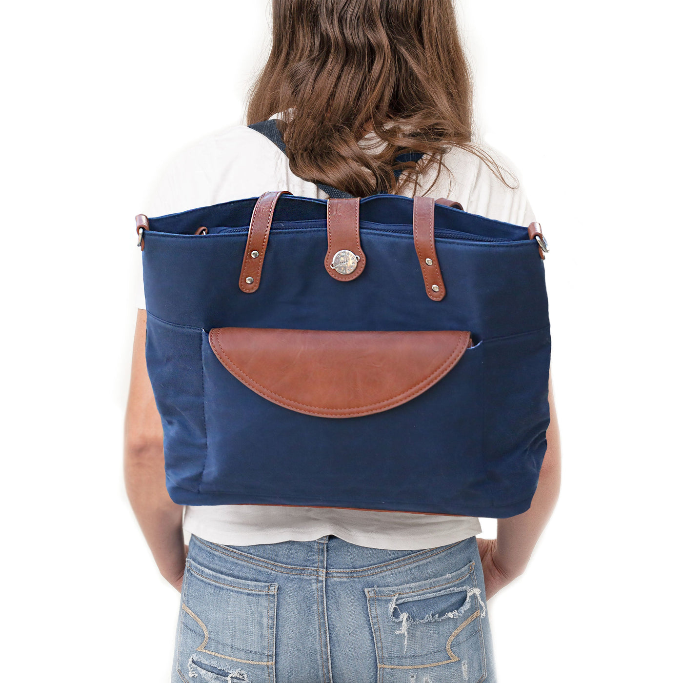 Mom in jeans and white t-shirt standing backwards wearing a navy blue waxed canvas tote bag as a backpack with brown vegan leather accents, all on a white background. 