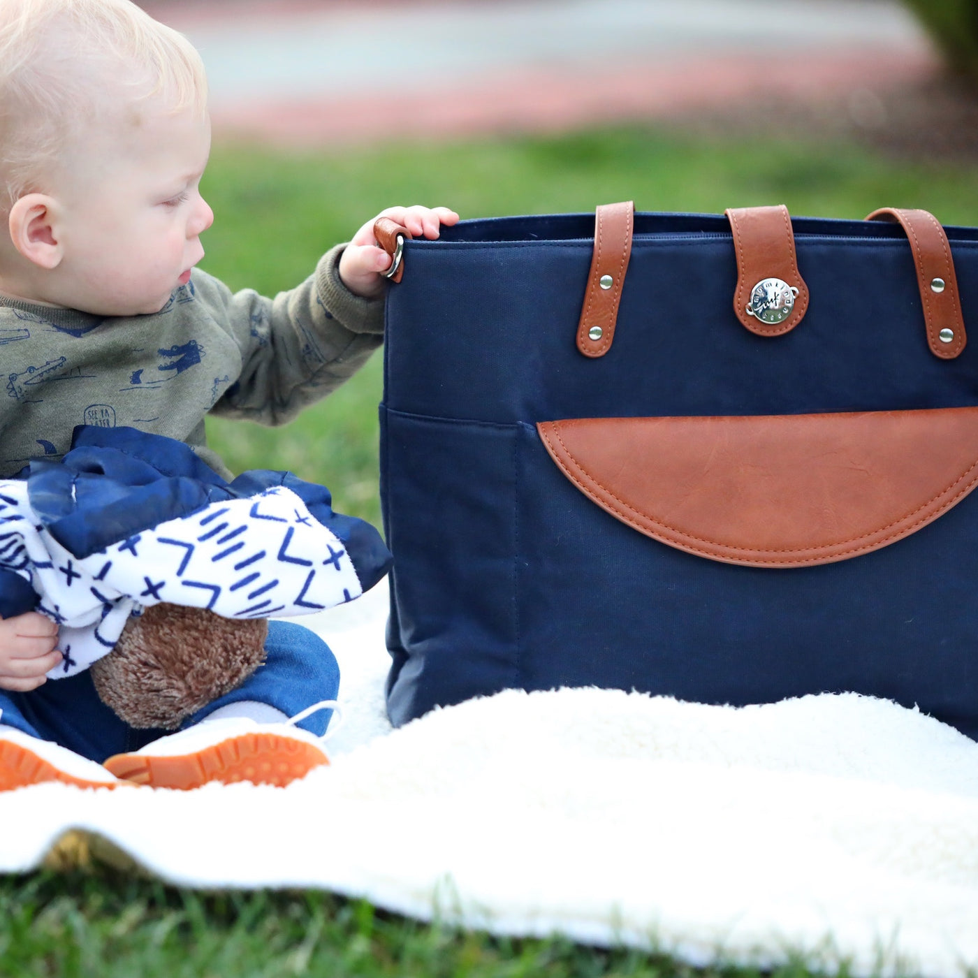 A baby sitting on a blanket outside on the grass, looking at a navy blue canvas tote which has caramel brown vegan leather accents.