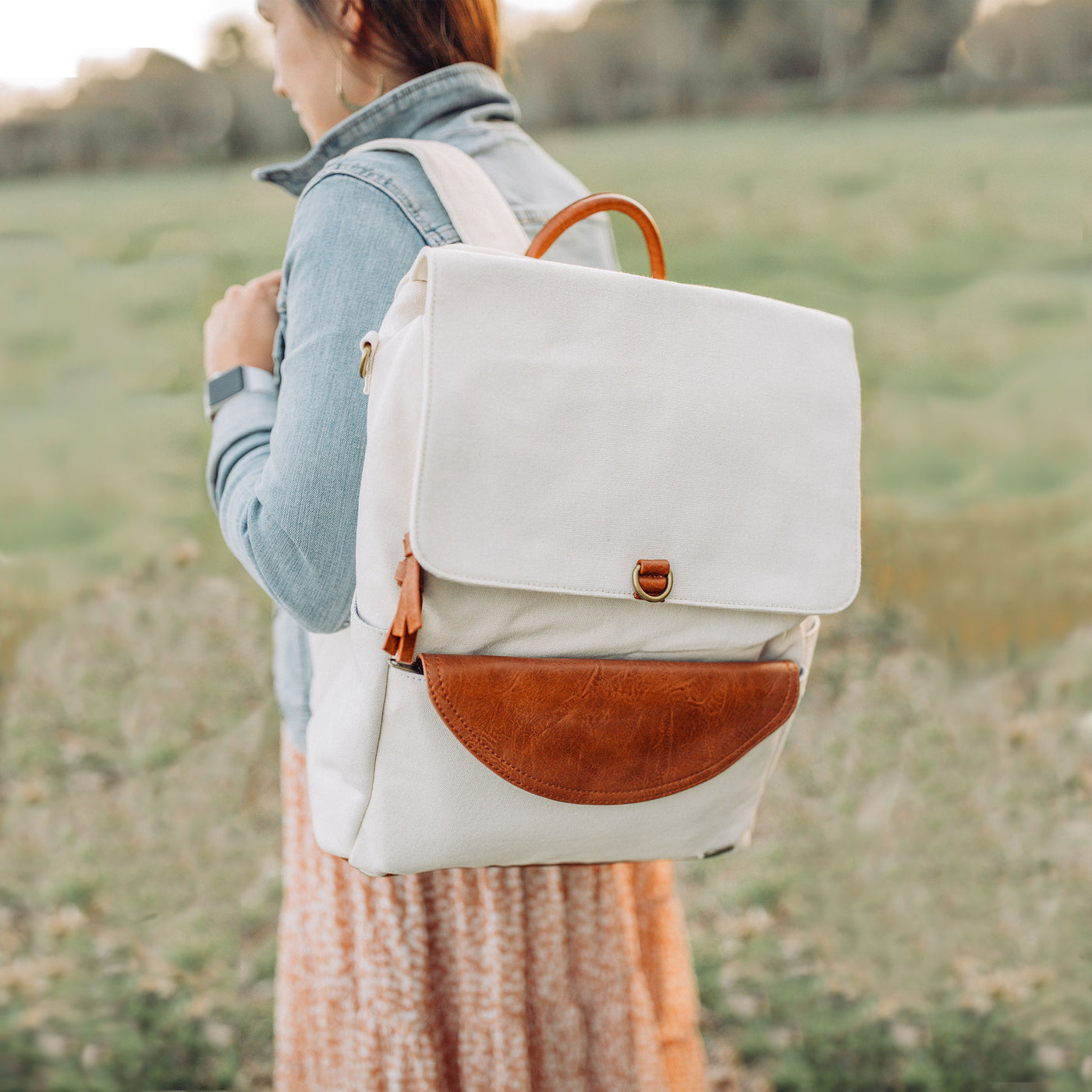 A woman standing sideways outside in a green field wearing a jean jacket and a stone colored canvas backpack with caramel brown vegan leather accents. 