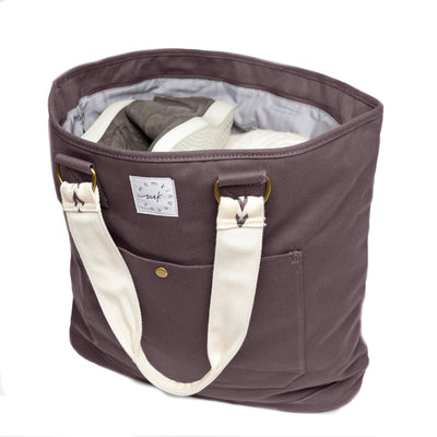 An extra-roomy canvas weekender tote in a graphite color with ivory embroidered shoulder straps with an arrow pattern, shown front-facing open and top-down filled with some clothing & shoes on a white background.