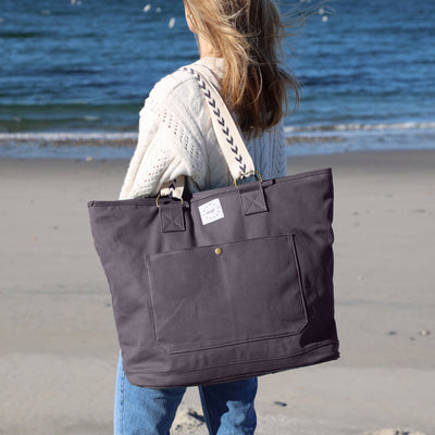 A woman standing on a beach wearing a graphic-colored canvas weekender tote on her shoulder with ivory embroidered straps featuring an arrow pattern. 