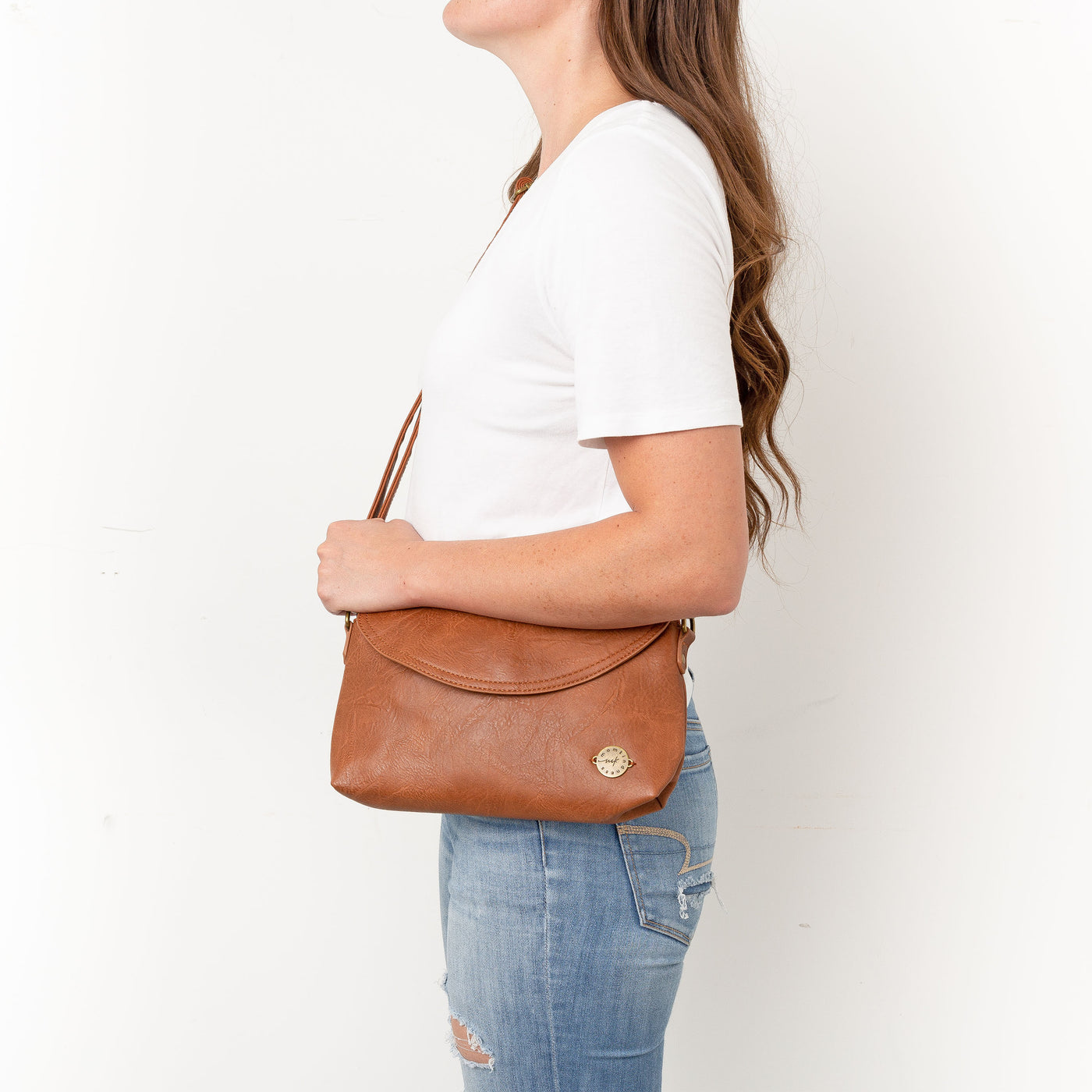A woman standing side-profile against a white background in jeans and white t-shirt wearing a caramel brown vegan leather crossbody clutch bag.