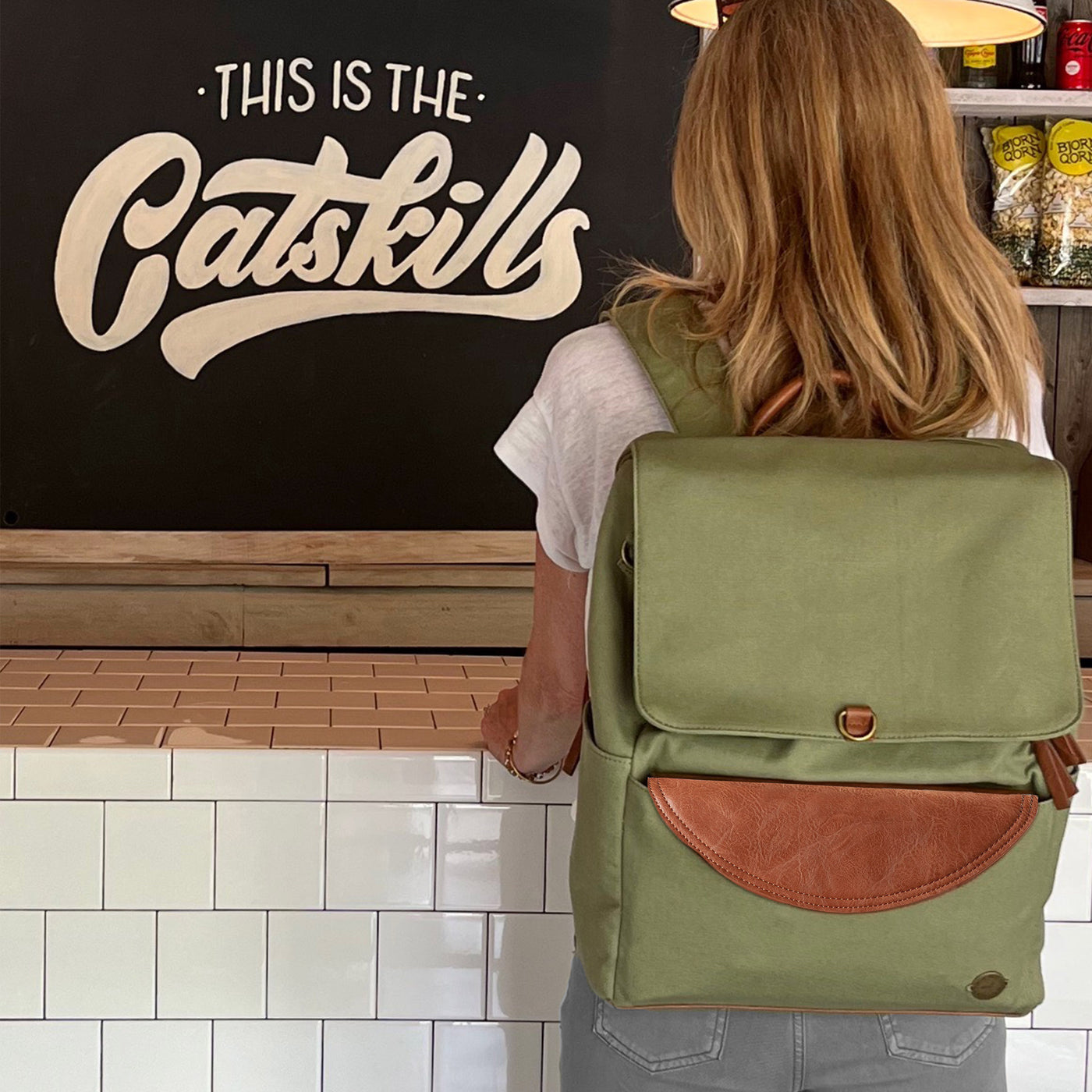 A woman wearing a laurel colored canvas everyday backpack with caramel brown vegan leather accents, standing in a small shop.