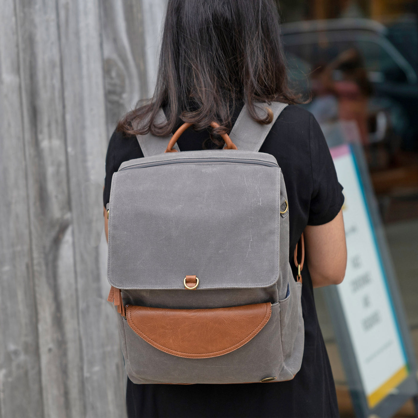 A woman standing backwards in front of a storefront wearing a waxed canvas grey backpack with caramel brown vegan leather accents. 