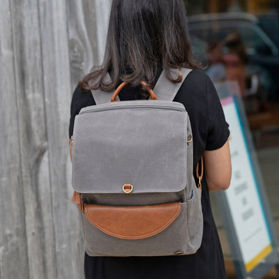 A woman standing backwards in front of a storefront wearing a luxurious grey backpack with caramel brown vegan leather accents. 