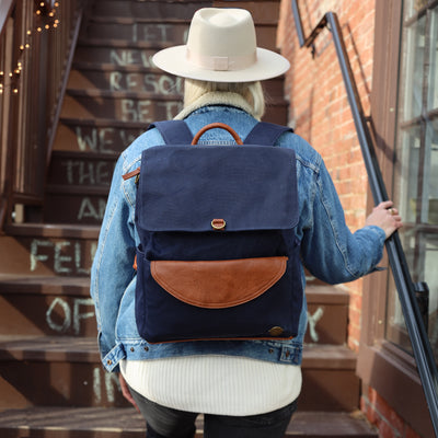 A woman in jean jacket and white hat walking up a brick outdoor stairway wearing a navy blue waxed canvas backpack with caramel brown vegan leather accents. 