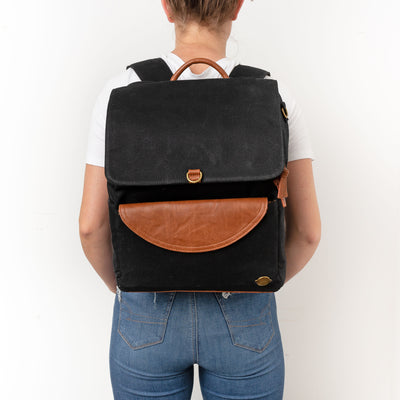 A woman standing backwards against a white background wearing a black canvas backpack with caramel brown vegan leather accents. 