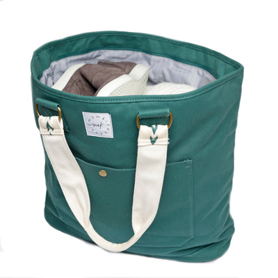 A large capacity canvas Juniper green colored tote shown at 3/4 angle and loaded with clothes, sneakers, etc.