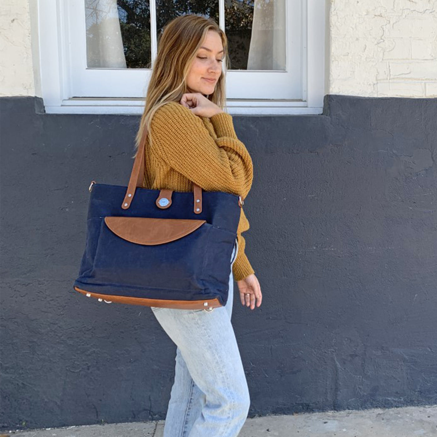 A woman in jeans and gold sweater carrying a navy waxed canvas carryall tote with caramel brown vegan leather accents on her shoulder, standing in front of a window background. 