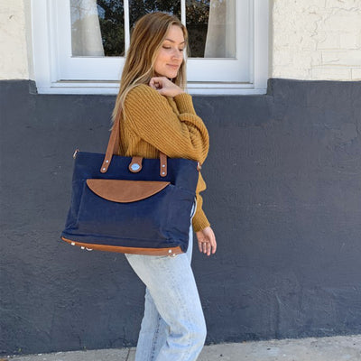 A woman in jeans and gold sweater carrying a navy waxed canvas carryall tote bag with caramel brown vegan leather accents on her shoulder, standing in front of a window background. 