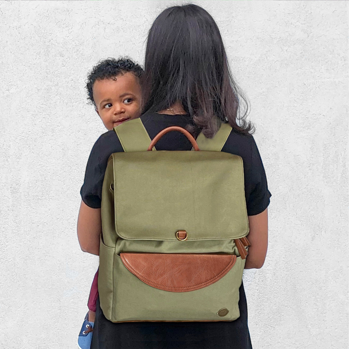 A woman holding her toddler son, wearing a laurel green colored canvas backpack diaper bag with brown vegan leather accents, standing backwards against a textured white wall. 