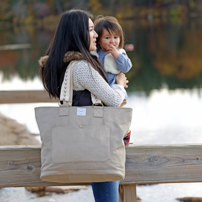 A mom wearing a rye (tan) color canvas tote with patterned shoulder straps while holding her toddler in an outdoor lake setting.