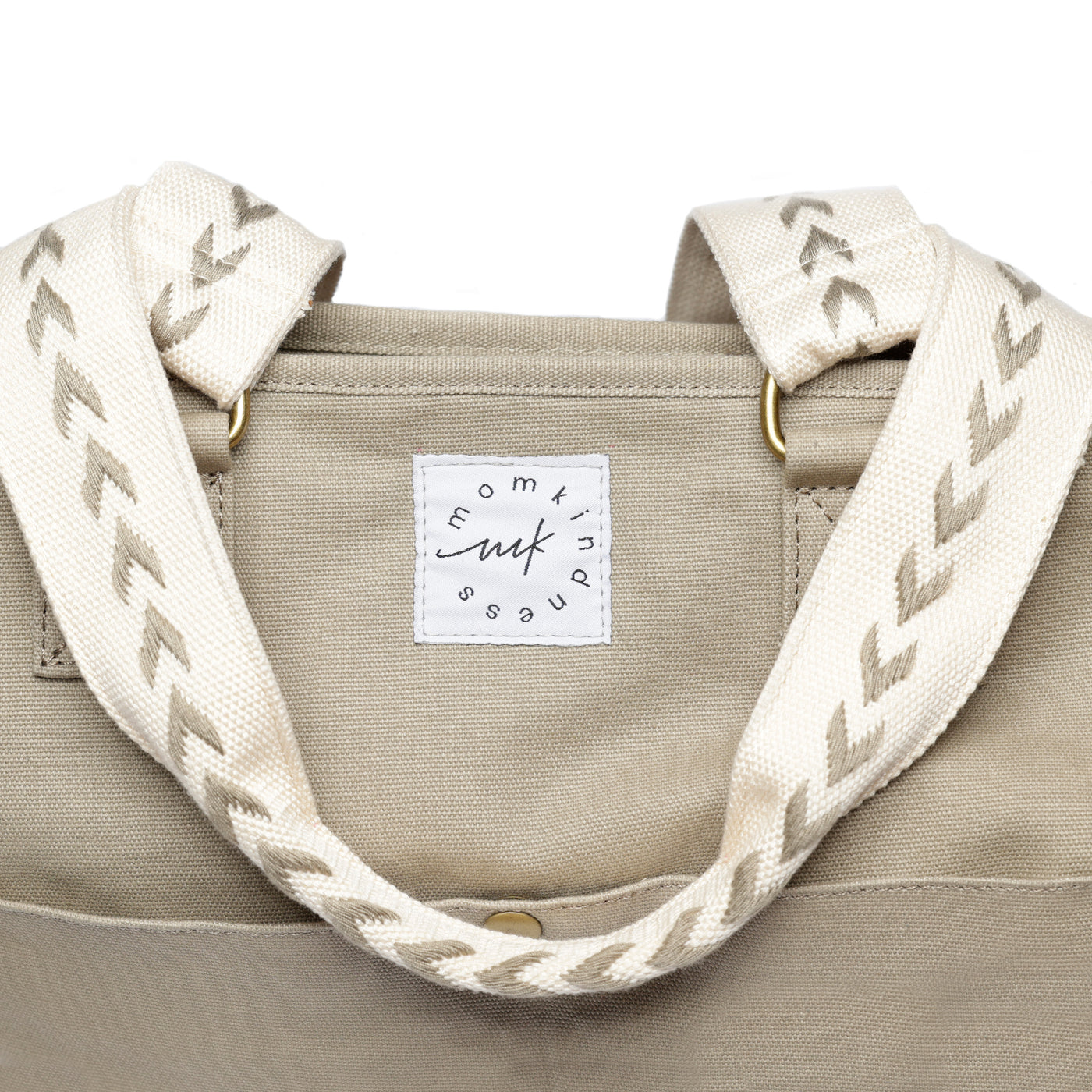 A close-up of the arrow pattern embroidered beige shoulder straps on a tan colored canvas tote and showing the Momkindness logo patch.