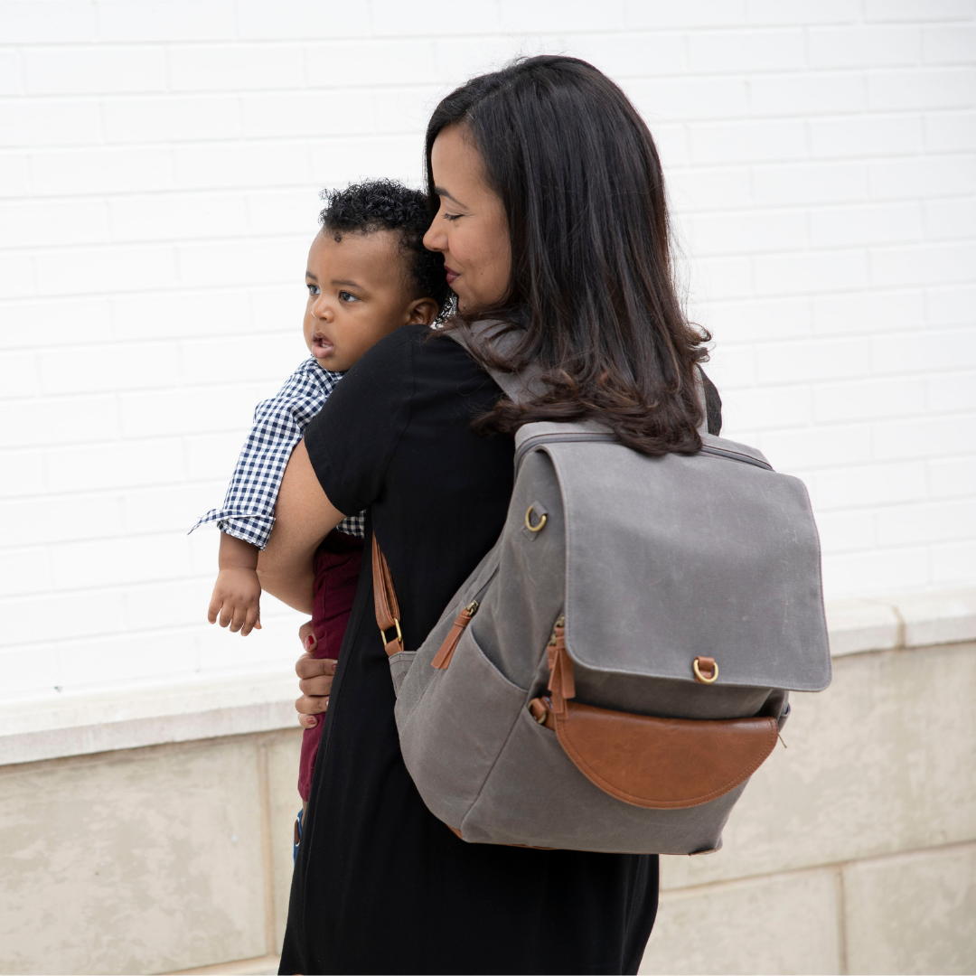 Women holding baby wearing a grey waxed canvas Duo Backpack with brown vegan leather accents on her back.  