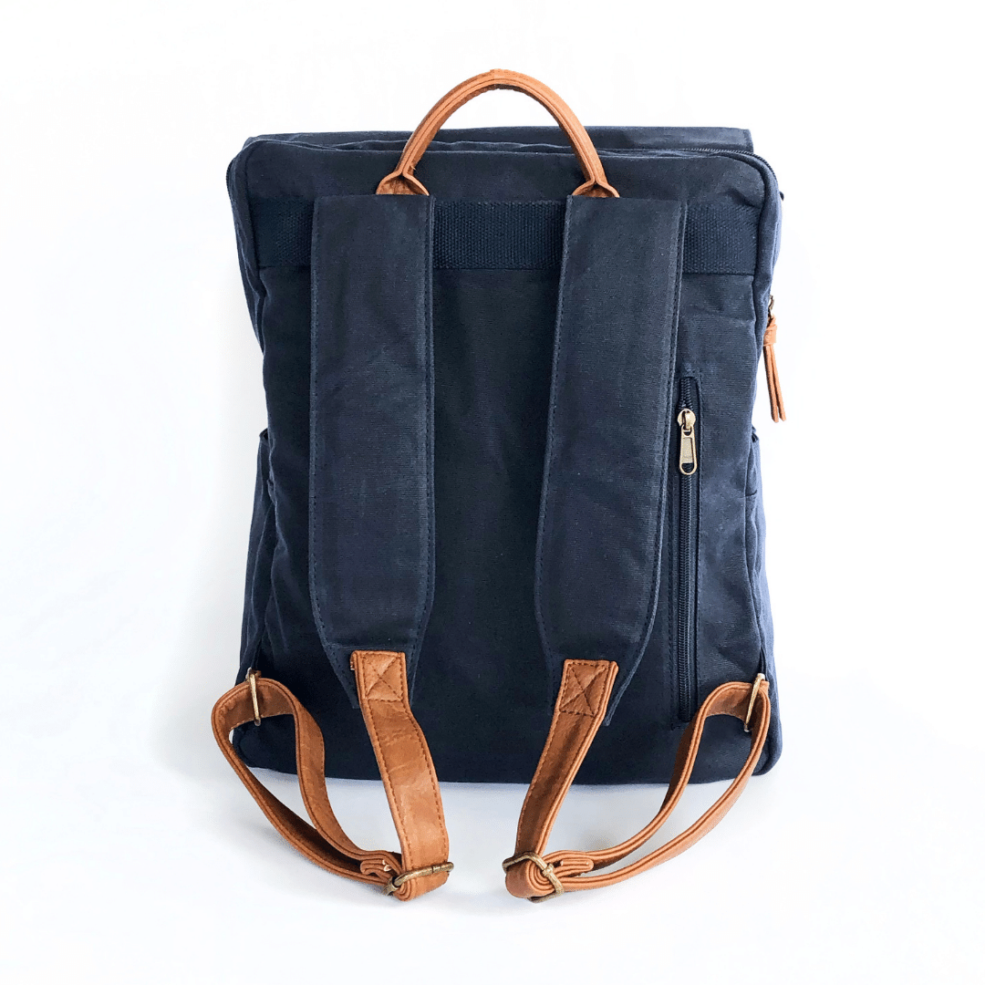 Backside view of navy waxed canvas backpack that shows extra comfort-padded canvas straps with brown vegan leather adjustable strap ends with bronze buckles and brown vegan leather carry handle on top. Image shown on a white background.