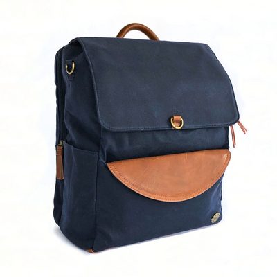 Front-facing 3/4 view of large capacity navy waxed canvas backpack with brown vegan leather accents and removable clutch. Side pocket and separate zip-top laptop pocket on back of pack can be seen as well as top carry handle. Image shown on white background.