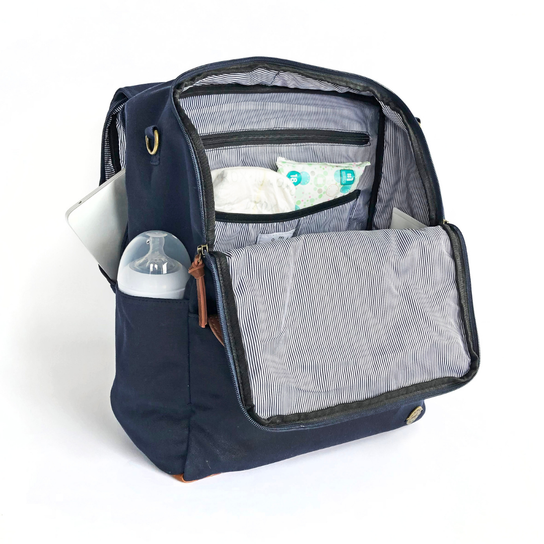 A front-facing 3/4 view of a navy blue waxed canvas backpack diaper bag with front flap zipped open showing pockets inside, a baby bottle in side pocket, and a laptop in separate back compartment. Wipe-clean lining is a thin black and white stripe pattern with black accents. Image shown on a white background.
