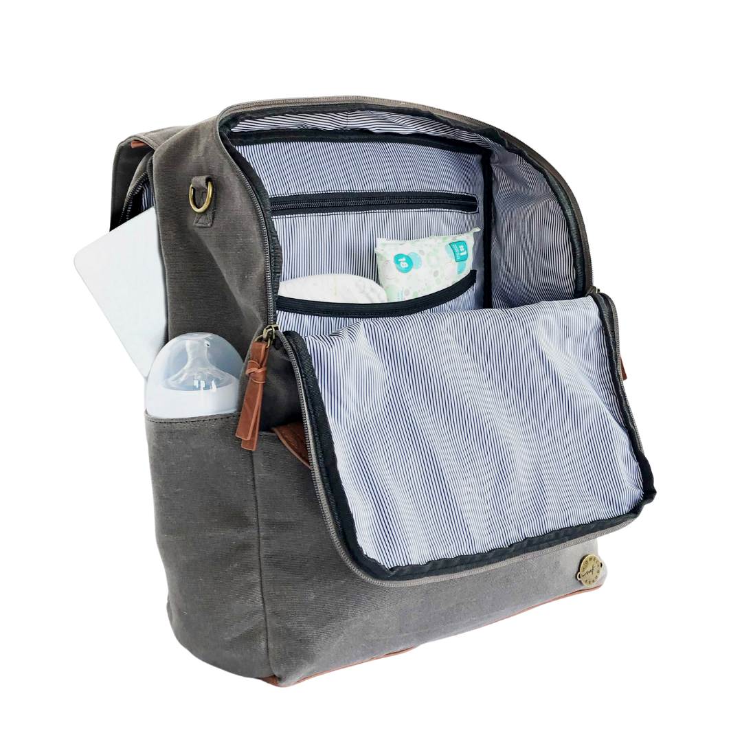 A front-facing 3/4 view of a grey waxed canvas backpack diaper bag with front flap zipped open showing pockets inside, a baby bottle in side pocket, and a laptop in separate back compartment. Wipe-clean lining is a thin black and white stripe pattern with black accents. Image shown on a white background.