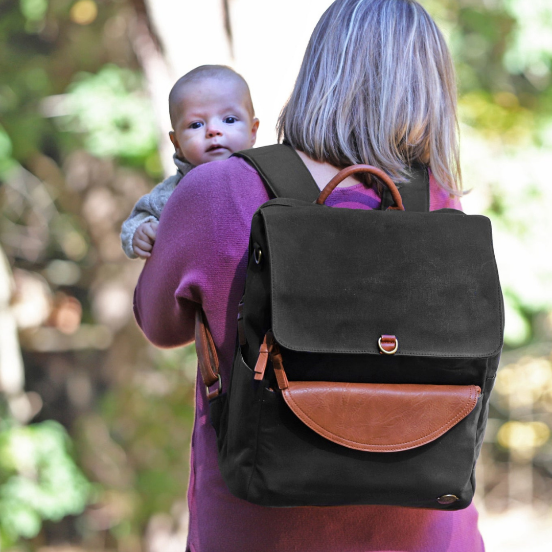 A mom in a pink sweater standing from behind while holding her baby peeking over her shoulder, walking on a forest path. Mom is wearing a black waxed canvas backpack with brown vegan leather accents.