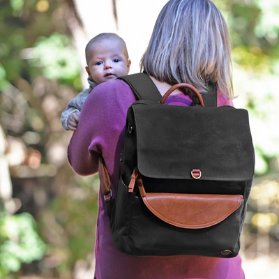 A mom in a pink sweater standing from behind while holding her baby peeking over her shoulder, walking on a forest path. Mom is wearing a black waxed canvas backpack with brown vegan leather accents.