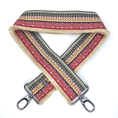 Close-up on white background of an adjustable length, woven bag strap with cream, red and green pattern with fringe on one side, and silver clasp.