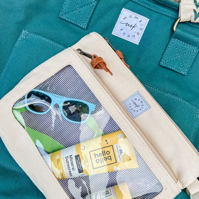 Close-up image of an ivory, stone-colored canvas packing zipper pouch with a clear view front pocket for of baby accessory items, laying on top of a juniper colored canvas tote.
